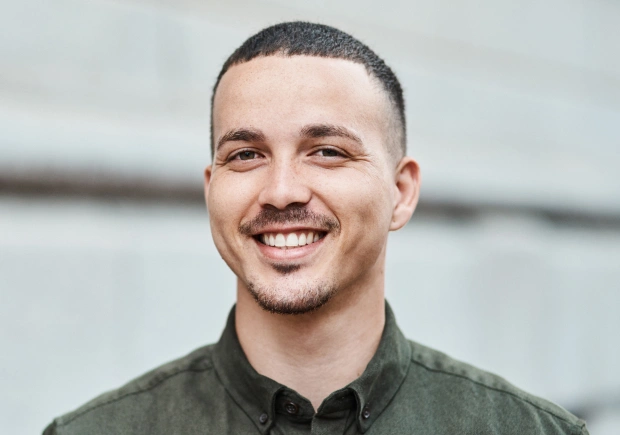 Man smiling at camera wearing olive green shirt against blurred white background.