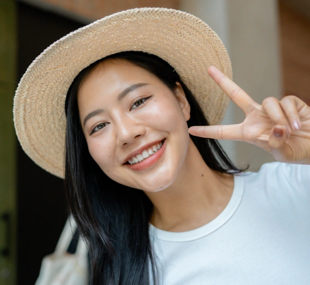Young woman wearing straw hat smiling at camera, adjusting hat brim indoors.
