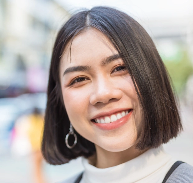 Young woman smiling at camera outdoors, wearing hoop earrings and light-colored top.