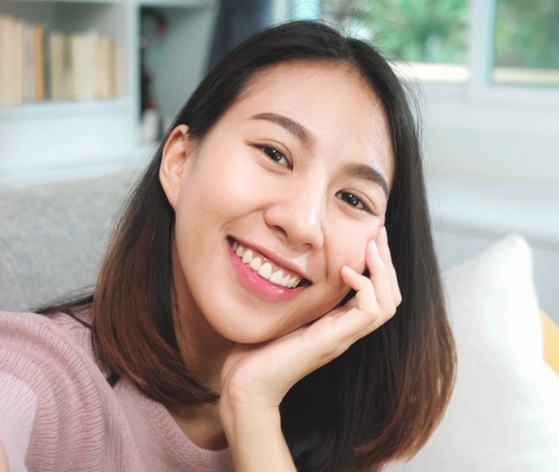 Young woman smiling at camera, hand on face, sitting on couch in bright room.