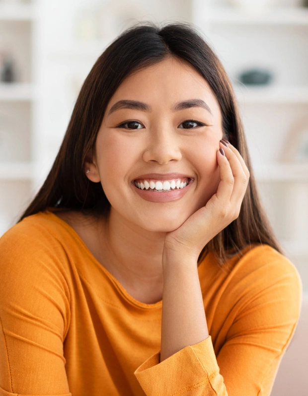 Young woman smiling cheerfully in orange shirt, hand on face, bright indoor setting.