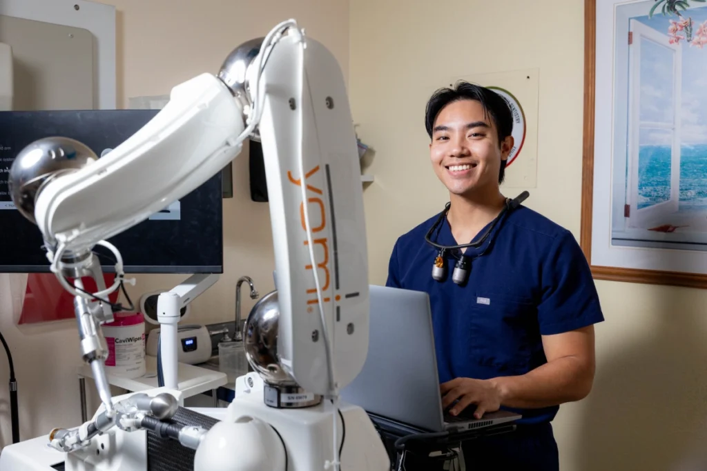 Dental professional in blue scrubs operates surgical microscope in clinical treatment room.