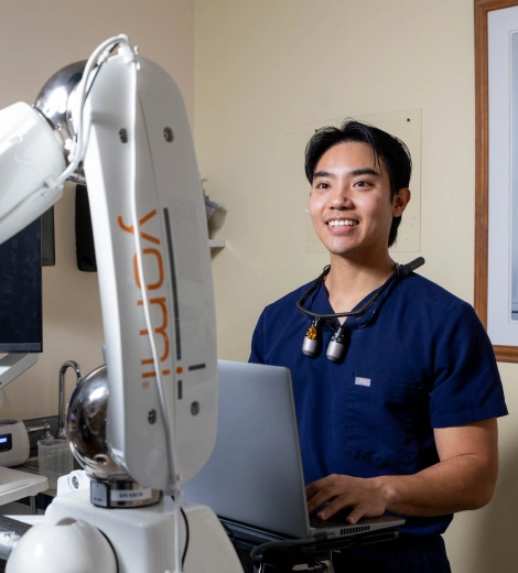 Dental professional in blue scrubs holding laptop next to dental equipment in clinical setting.
